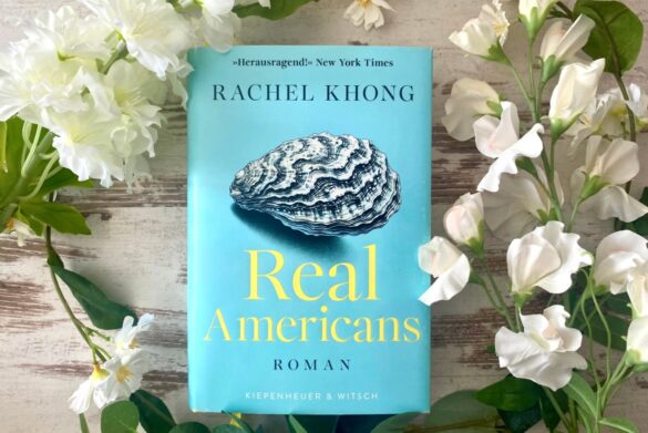 Buch „Real Americans“ von Rachel Khong auf hellem Holzboden mit dekorativen Blumen arrangiert – minimalistische Buchinszenierung für Rezension