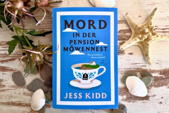 Beitragsbild zur Rezension von Mord in der Pension Möwennest von Jess Kidd. Das Buch liegt auf einem hellen Holzboden umrahmt von Muscheln und einem Seestern